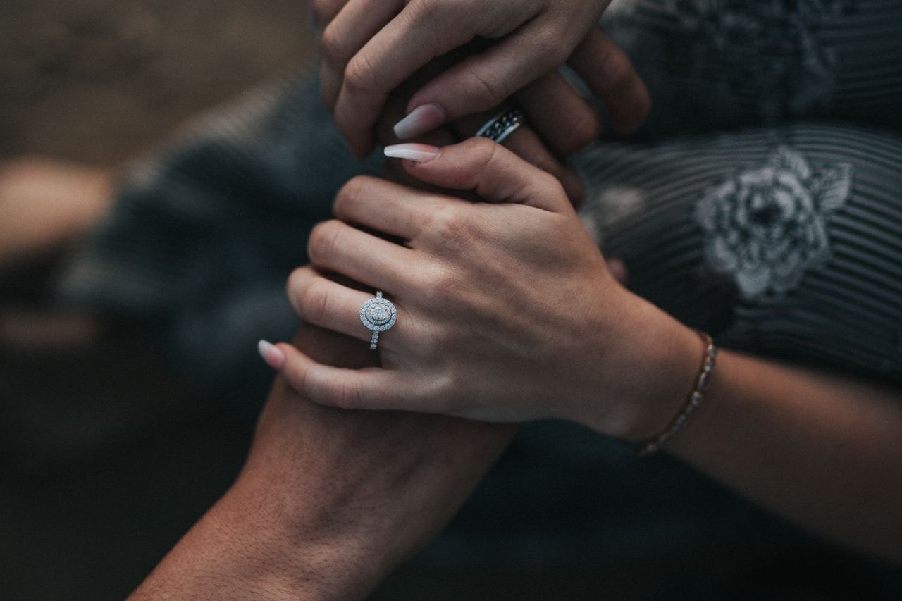 close up image of a couple holding hands, the woman wearing a halo and side stone oval cut engagement ring 