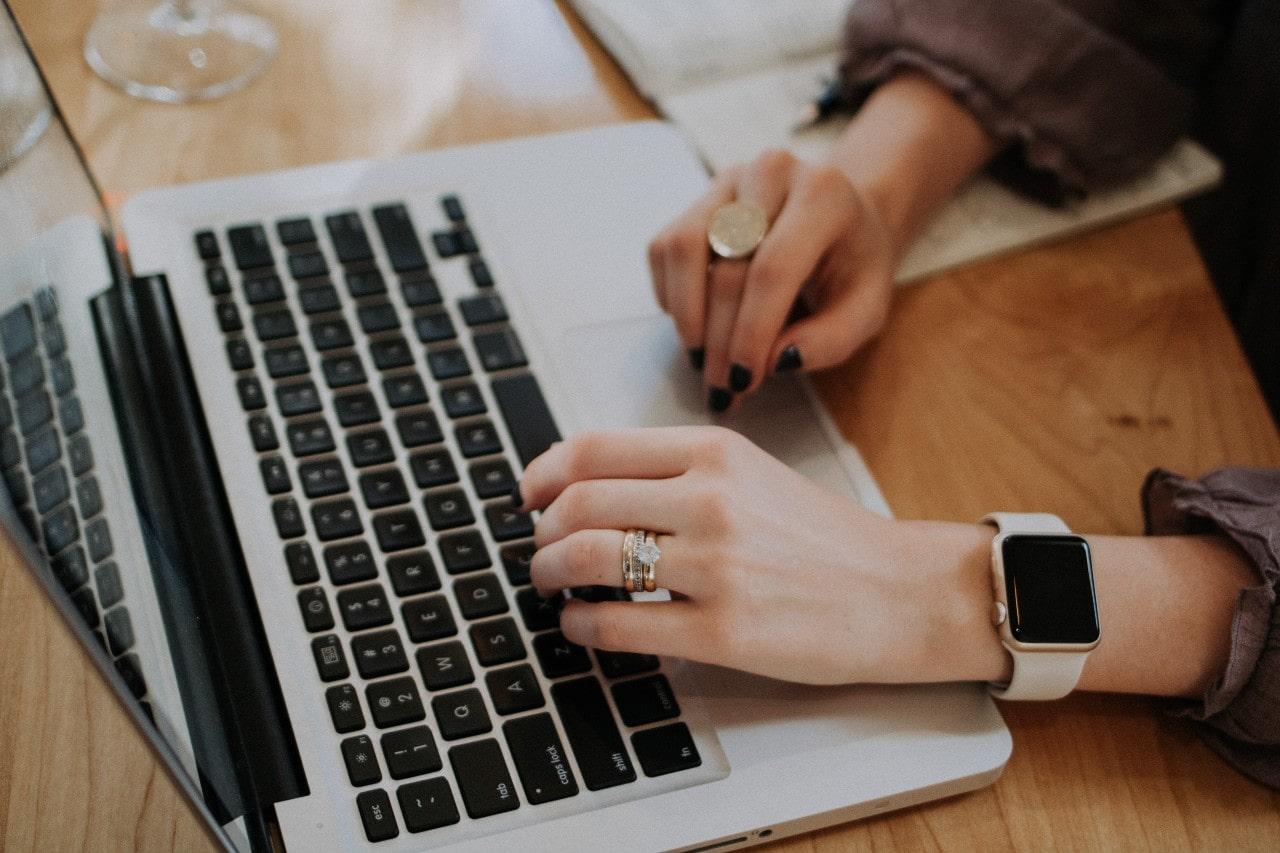 An older, married woman sips wine while browsing jewelry on her Macbook in her kitchen