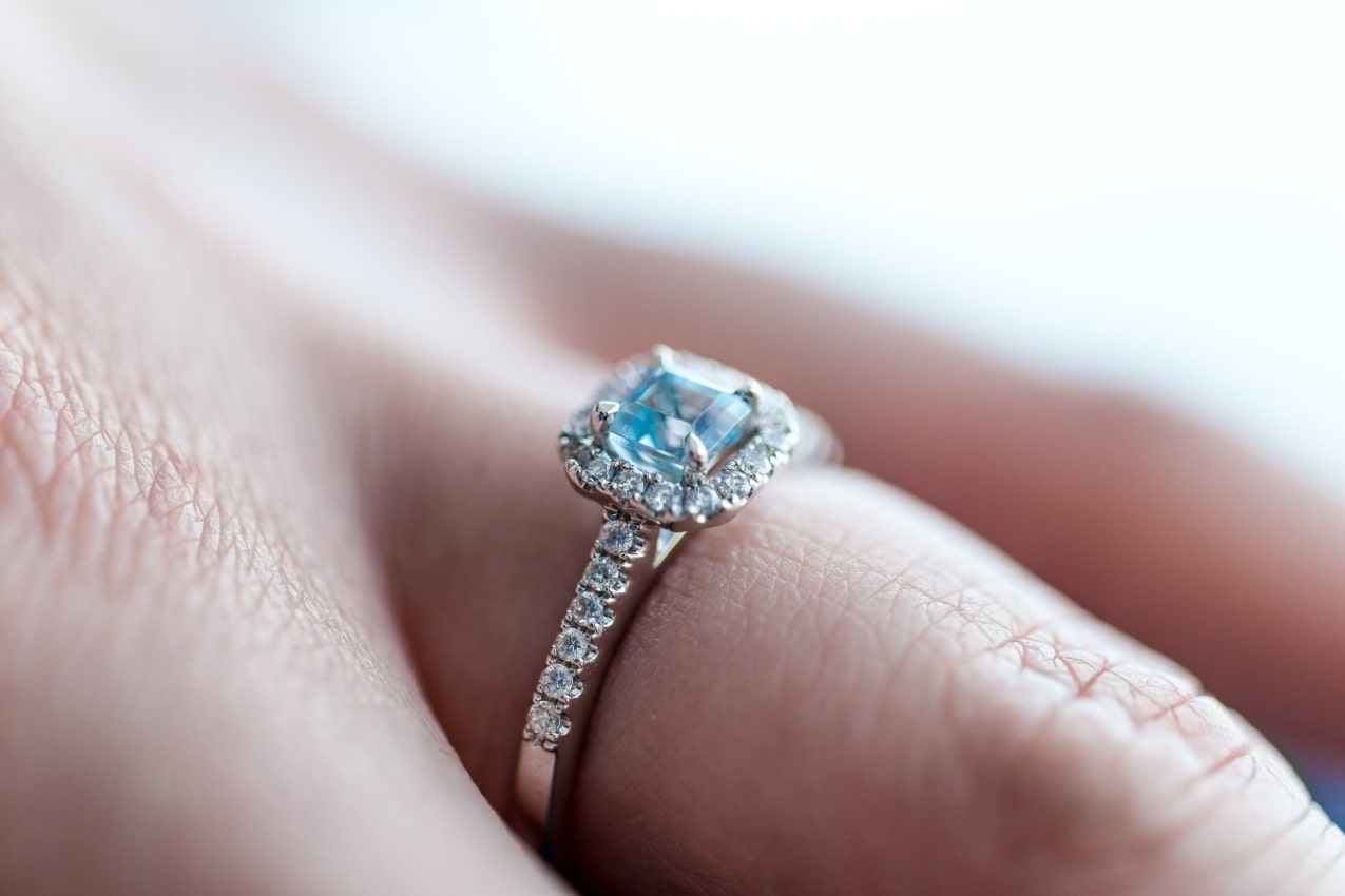 Close-up image of a hand wearing a princess cut engagement ring with a blue center stone