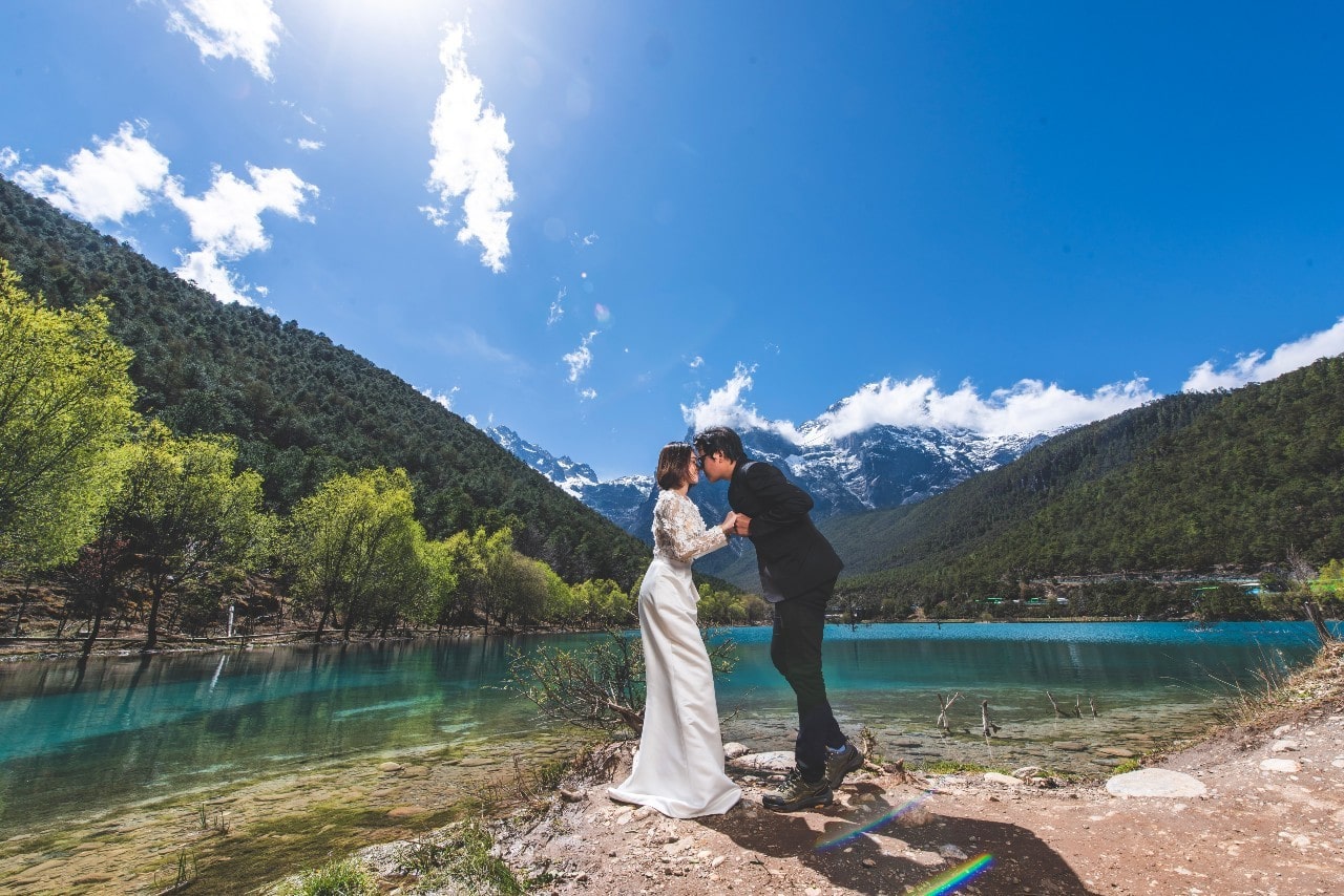 a couple embracing by a body of water in the shadow of green mountains