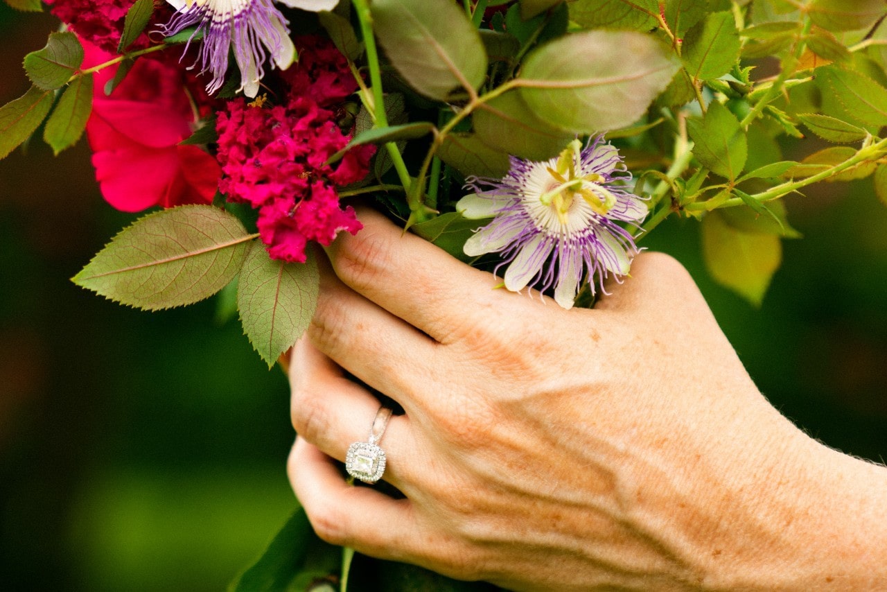 a woman’s hand holding a bouquet of flowers and wearing a princess cut engagement ring