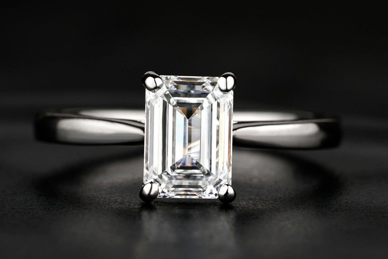 A close-up of a simple yet mesmerizing solitaire ring with an emerald cut center stone on a black backdrop.