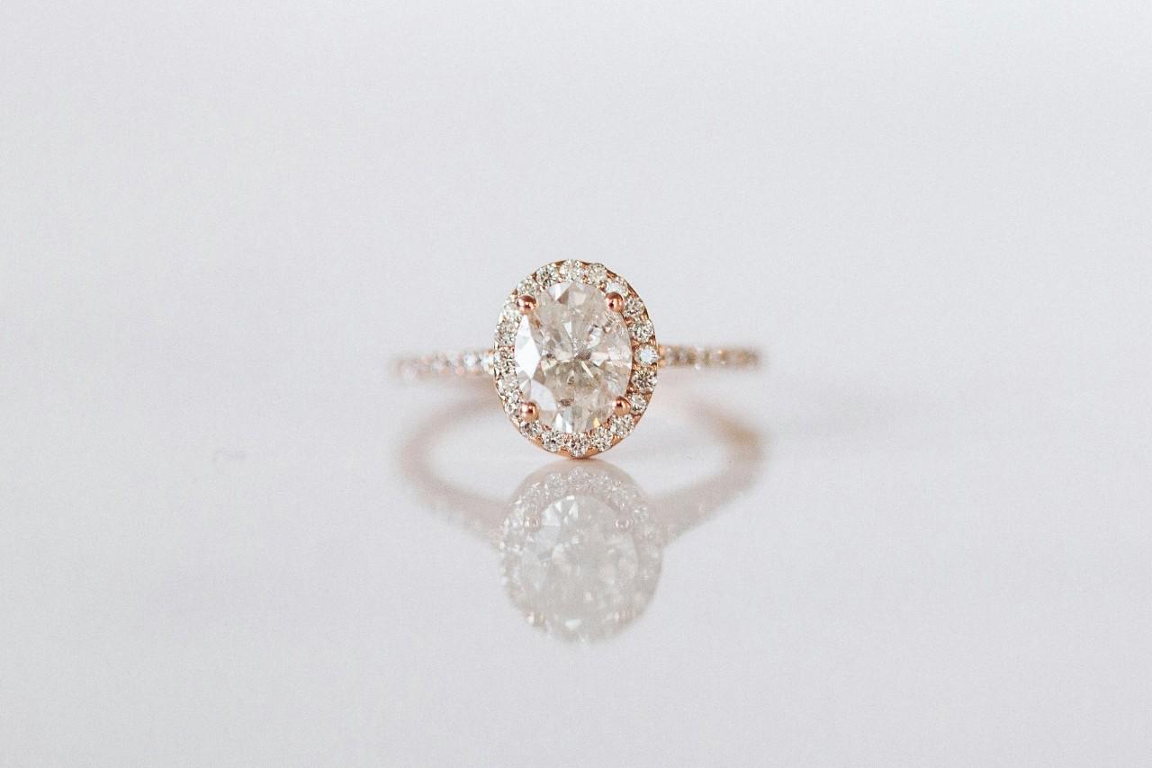 A rose gold halo engagement ring with an oval cut center stone displayed on a reflective white background.