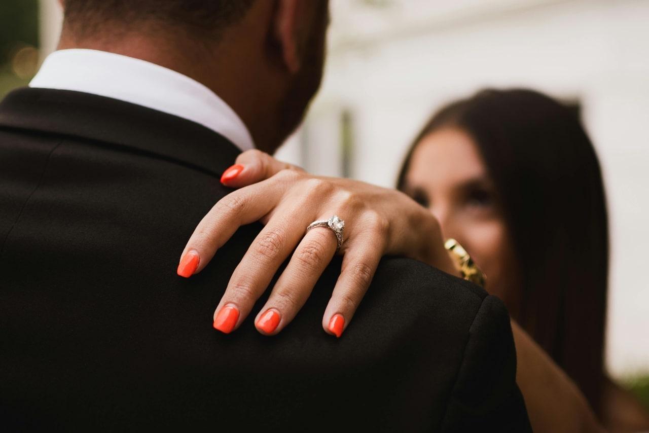 A bride’s hand resting on her groom’s shoulder, a white gold engagement ring on her finger.