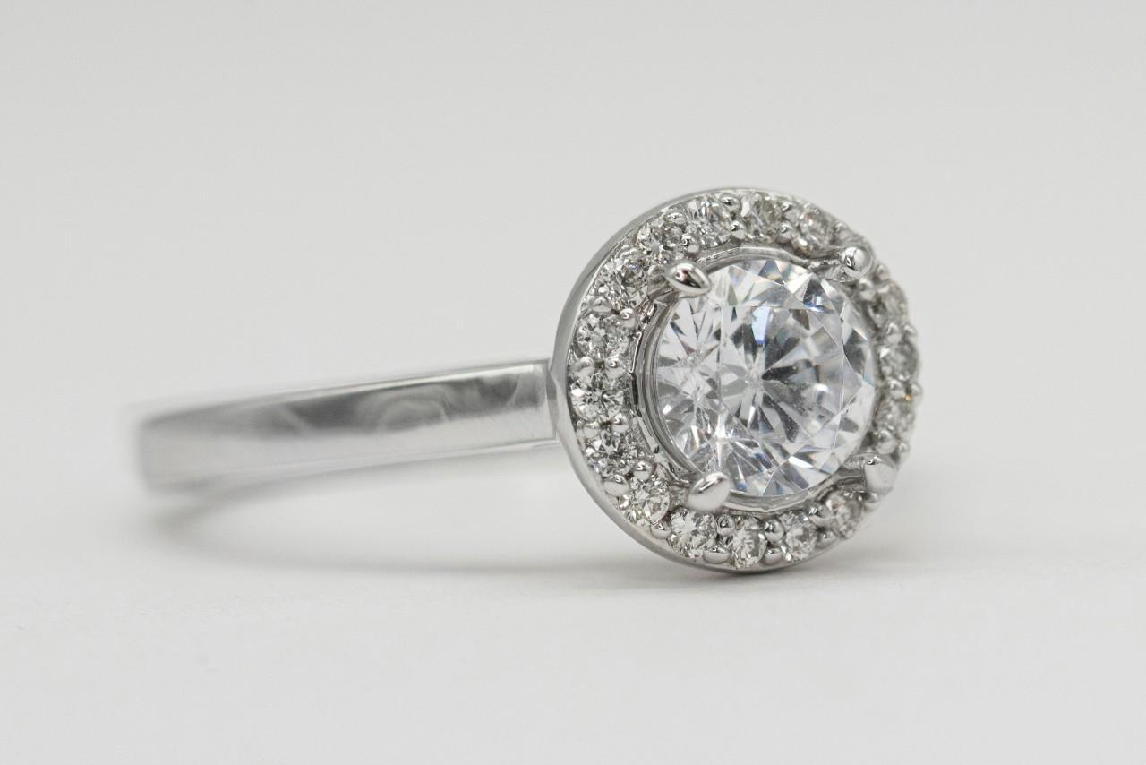 A close-up of a white gold bezel set halo engagement ring on a white surface.