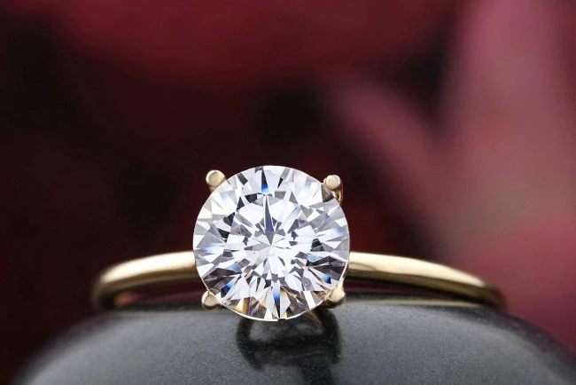A close up of a yellow gold solitaire round cut diamond engagement ring against a blurry burgundy background