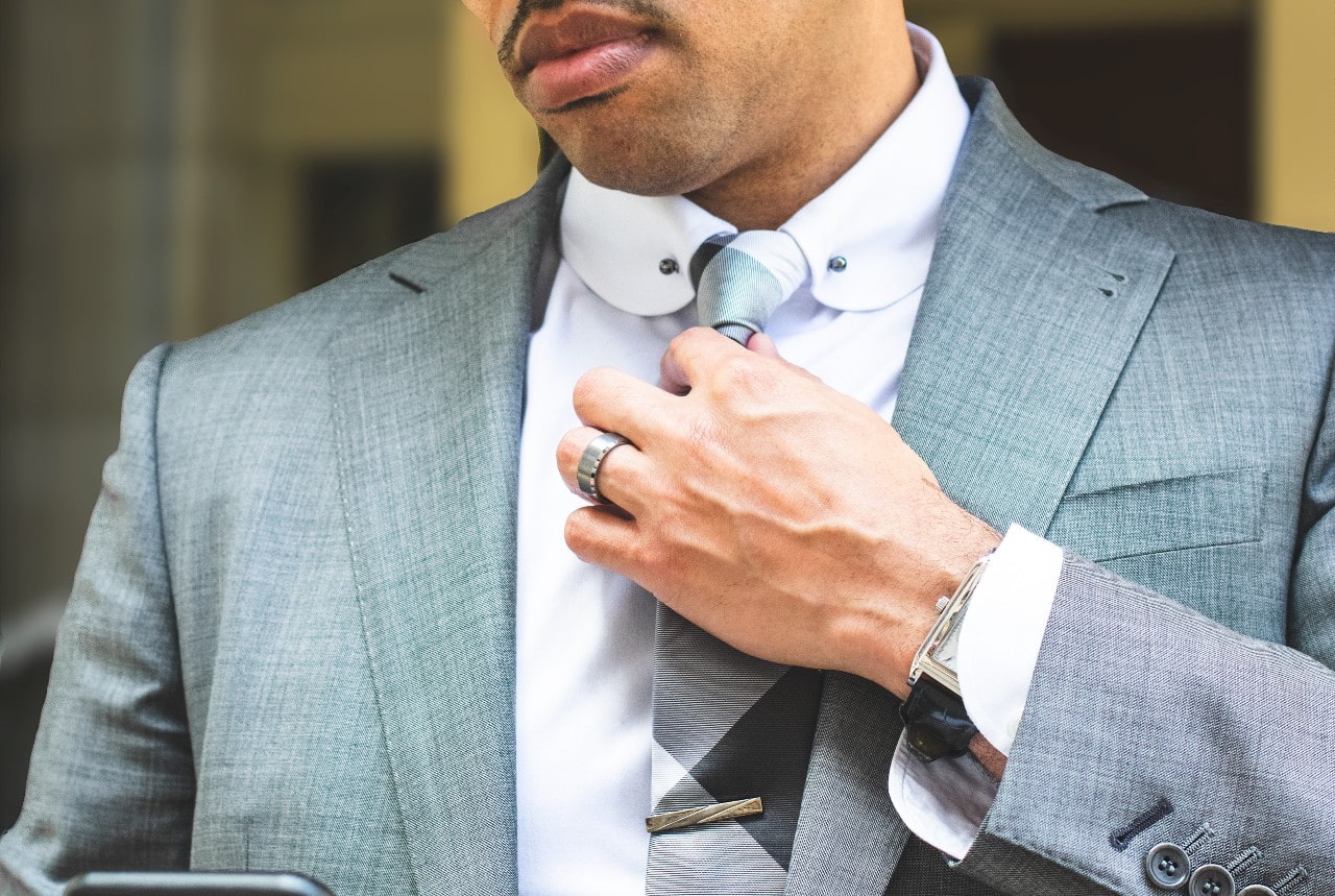 A close up of a guy in a gray suit fixing his tie with a silver wedding band.