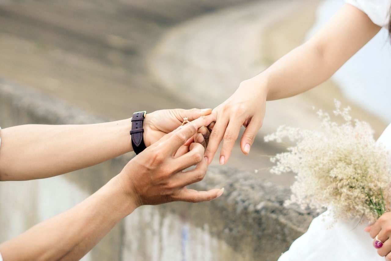 a person placing an engagement ring on a woman’s hand who is wearing white and holding flowers