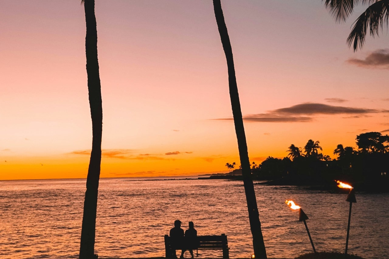 a couple sitting on a bench by the sea in Hawaii next to tiki torches as sunset