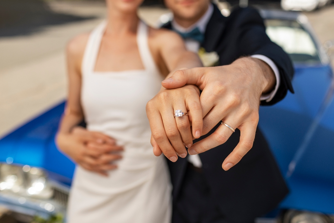 A newlywed couple leans on a retro deep blue car, showcasing a diamond engagement ring on a woman&rsquo;s hand. 