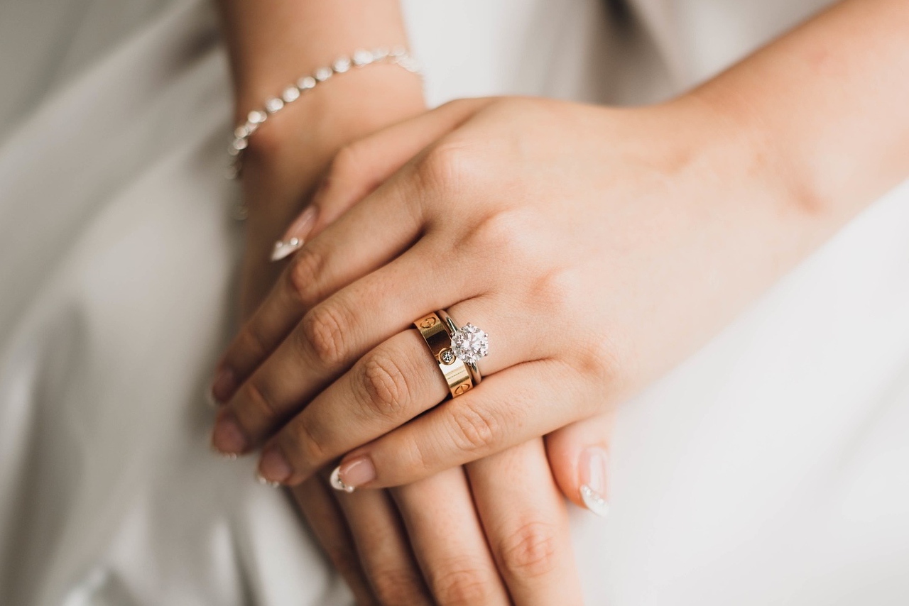 A brides hands with manicure nails on top of her wedding gown, showcasing a solitaire diamond engagement ring and yellow gold diamond wedding band