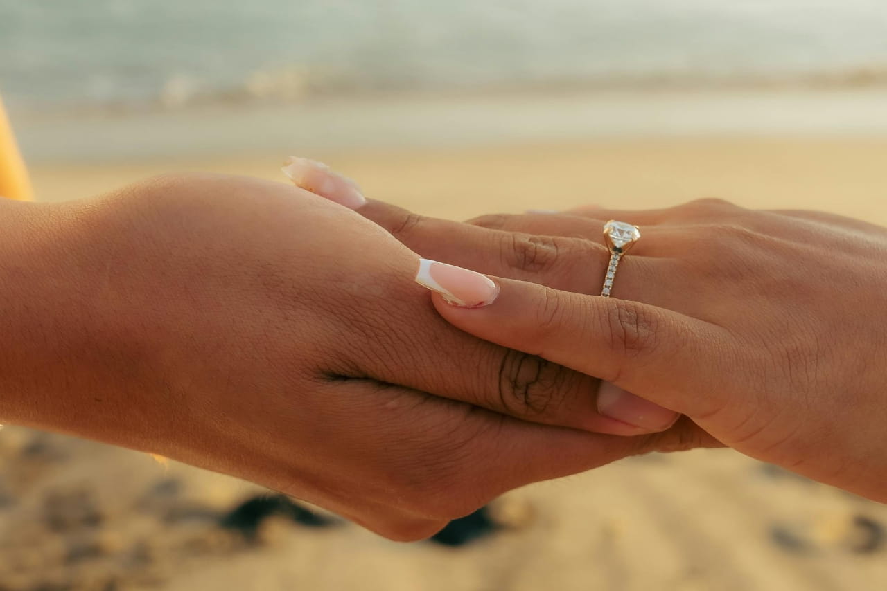 A hand with french manicure rests on another person&rsquo;s hand, showcasing side stone diamond engagement ring