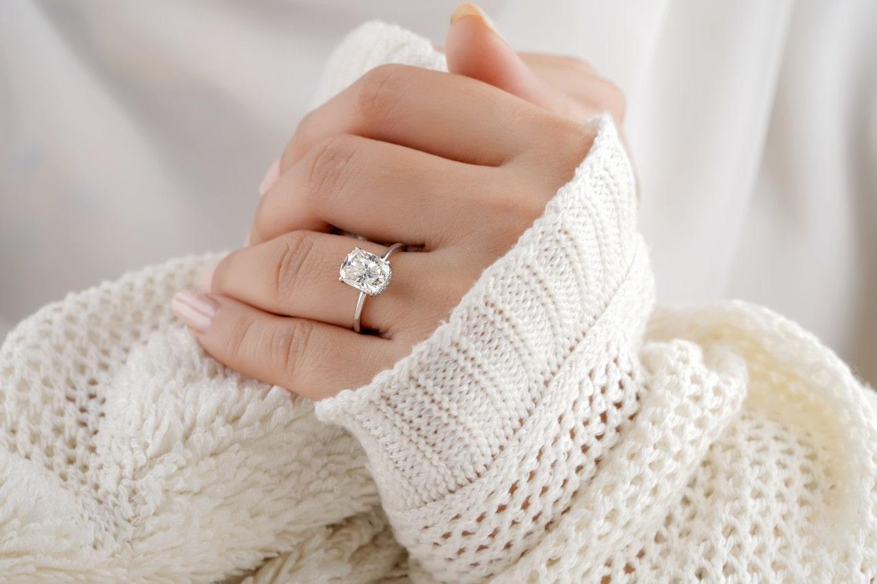 A woman’s hand wearing a silver, oval cut engagement ring and a white sweater.