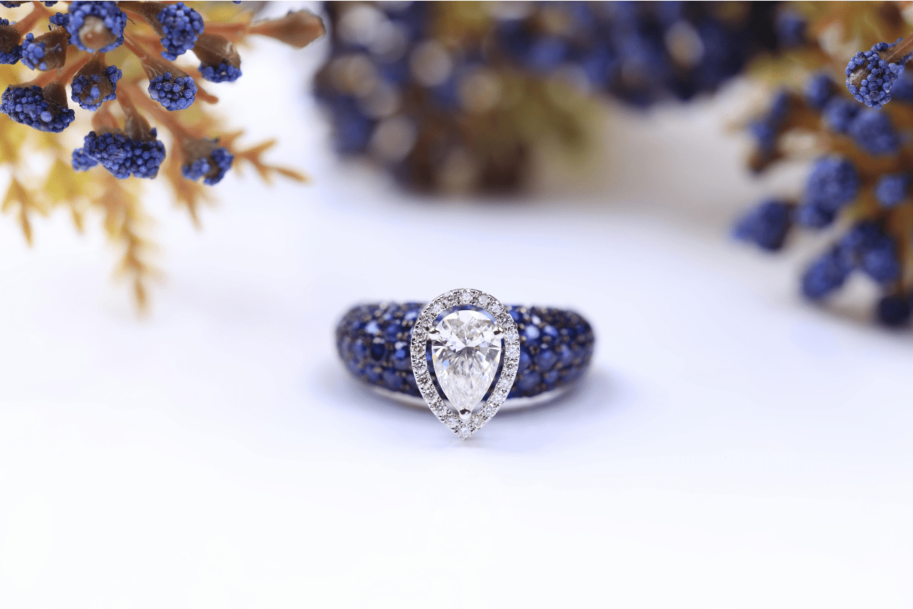 A pear cut halo engagement ring with pavé sapphire side stones displayed among sprigs of juniper berries. A pear cut halo engagement ring with pavé sapphire side stones displayed among sprigs of juniper berries.