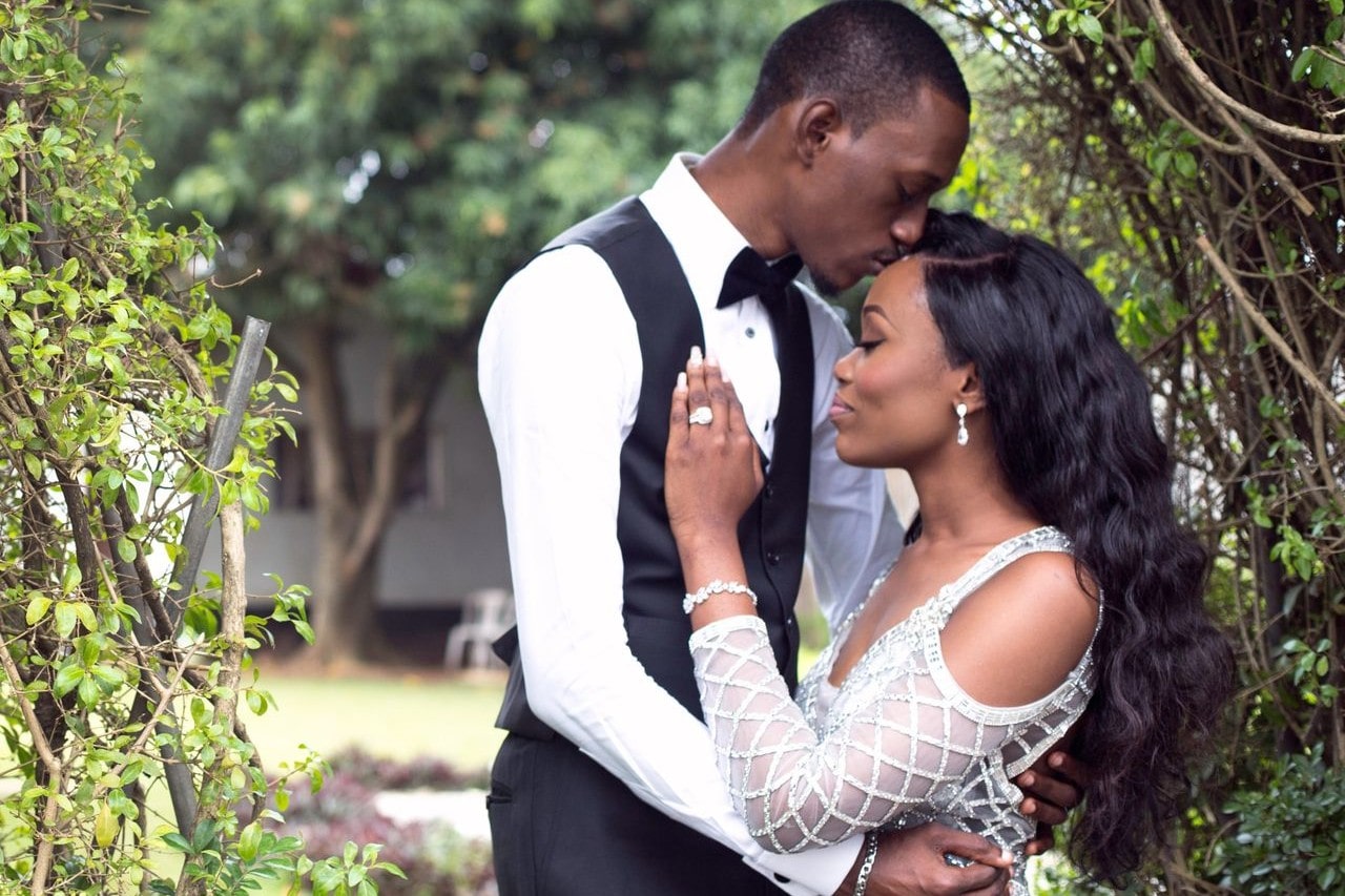 A bride and groom embrace by a fence outside.