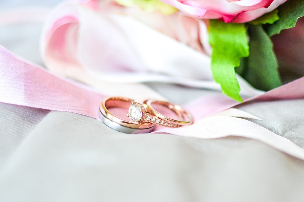 two rose gold wedding bands and a side stone engagement ring lying on a pink ribbon