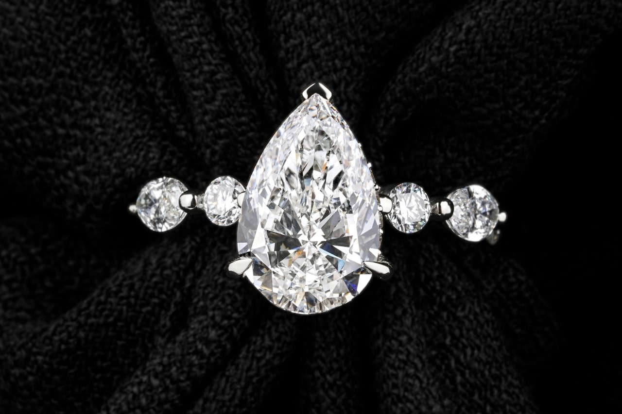 A close-up of a distinctive engagement ring with a pear center stone and large diamonds along the band. A close-up of a distinctive engagement ring with a pear center stone and large diamonds along the band.