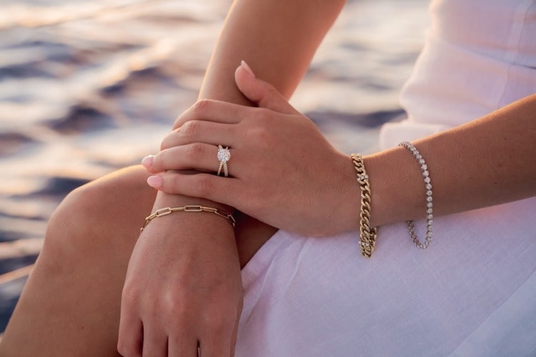 A woman sitting by the water at sunset wearing two yellow gold chain bracelets, a white gold diamond bracelet, and a split shank engagement ring