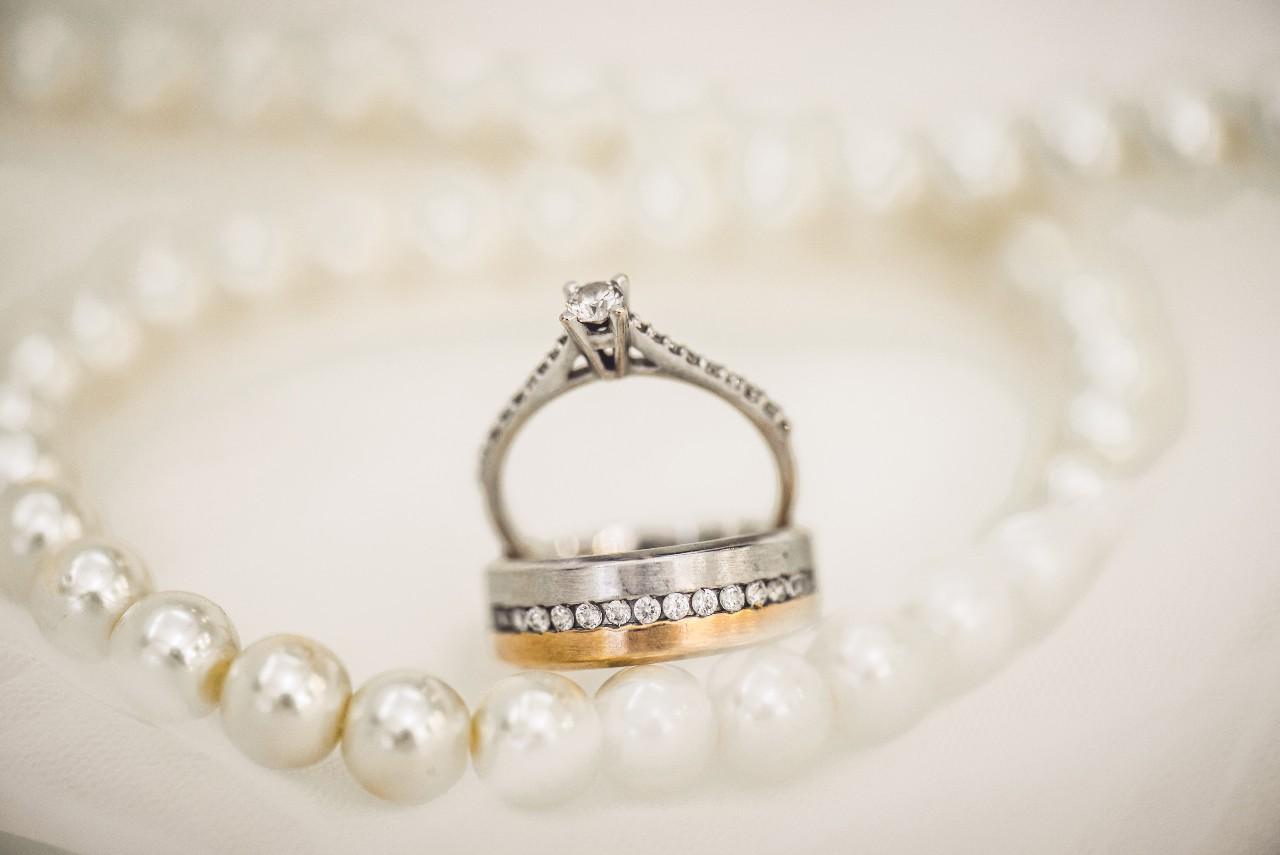 An engagement ring and a wedding band sit among a pearl necklace on a white cloth.