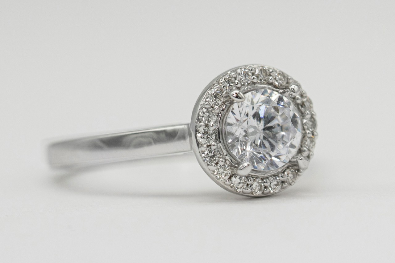 A close-up of a white gold bezel set halo engagement ring on a white surface.