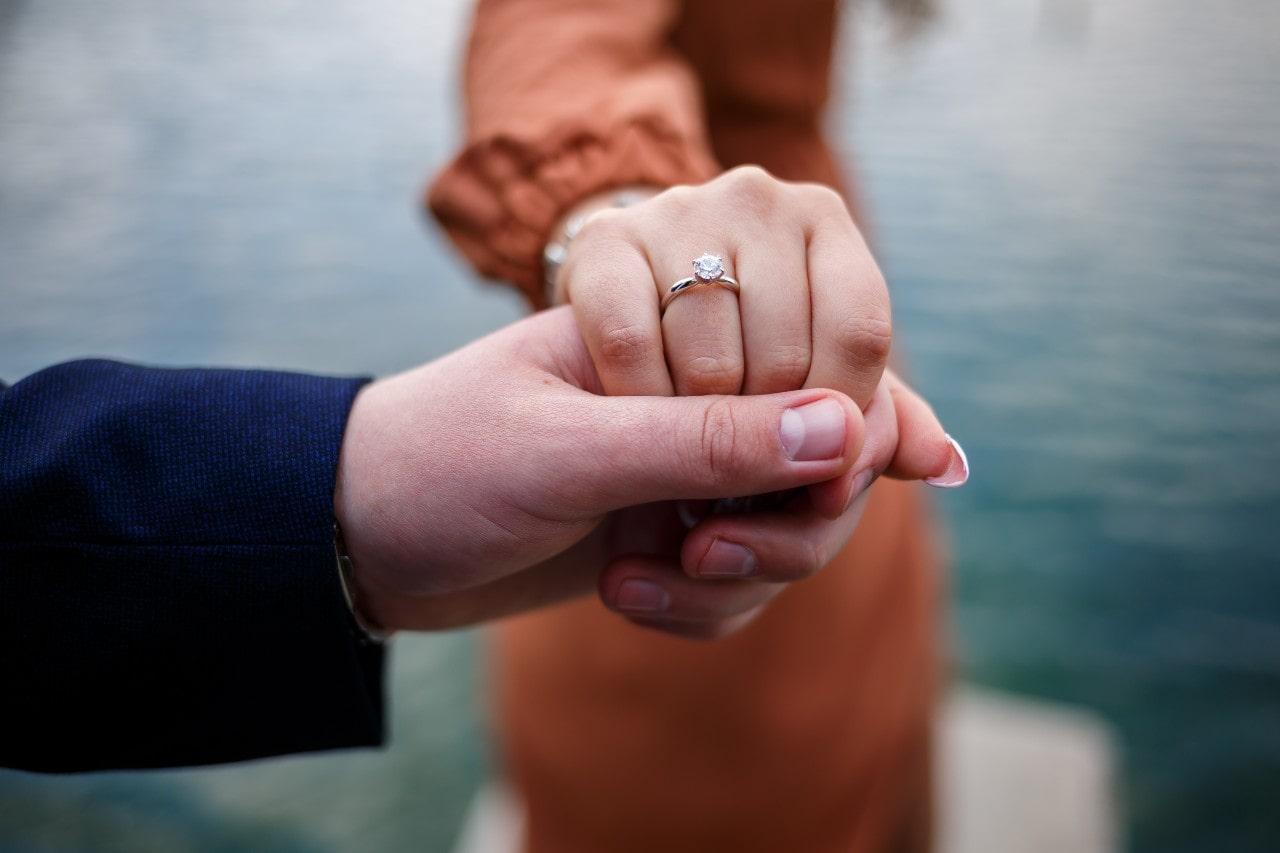 Close up on the hand of a woman wearing a solitaire ring holding her soon-to-be spouses hand next to a dock beside a body of water
