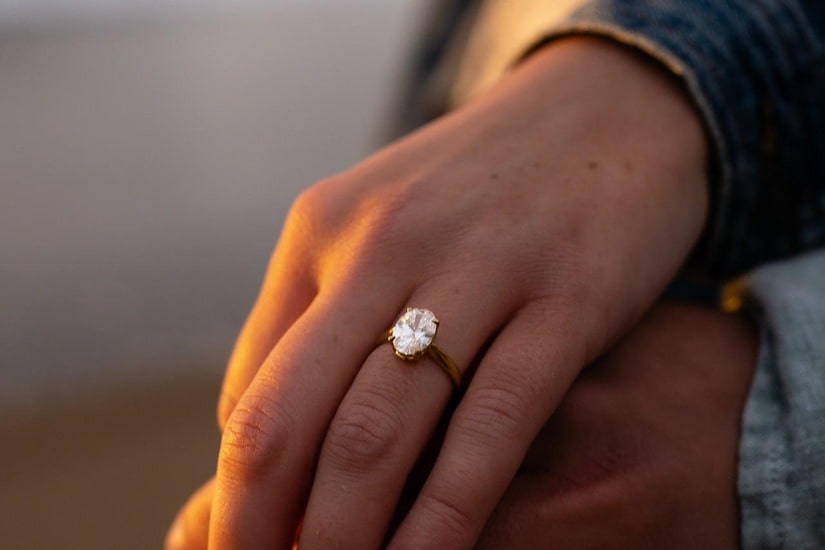 Close up on an oval cut engagement ring on the hand of a woman holding hands with her love at sunset on the beach.